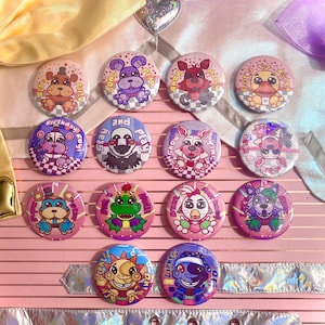 Puede incluir: Una colección de botones redondos y coloridos con personajes de dibujos animados y frases como "Let's Party" y "Stay and Play". Los botones tienen un acabado brillante y están dispuestos sobre una superficie rosa.