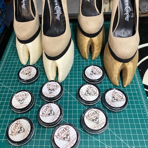 Könnte beinhalten: Beigefarbene High Heels mit hufförmigen Plateaus, beschriftet mit "Creature Cosplay". Mehrere kleine, runde Behälter mit dem Text "REAPERS" und einer Schädelgrafik sind ebenfalls sichtbar. Die Artikel sind auf einer grünen Rasteroberfläche angeordnet.