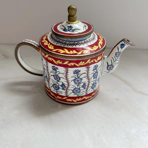 Puede incluir: Una tetera decorativa con base blanca, adornada con motivos florales azules y detalles rojos y dorados. La tapa presenta un pomo de latón y un diseño floral a juego. El asa y el pico también están decorados con motivos florales.