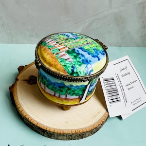 May include: A small, round, porcelain trinket box with a painted scene of a forest. The box has a gold trim and sits on a wooden base. The box is labeled "E50013-30 STAMP HOLDER" and "making Tradition Received".