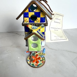 Könnte beinhalten: Ein dekoratives, dreistöckiges Vogelhaus mit einem skurrilen Design. Das Vogelhaus hat ein blau-gelb kariertes Dach, ein grünes Vogelhaus und eine Basis mit einer Blume und einer Biene. Am Vogelhaus ist ein Echtheitszertifikat angebracht.