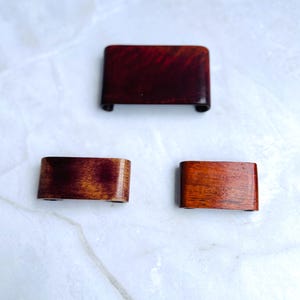 Könnte beinhalten: Drei kleine, gebogene Holzobjekte in verschiedenen Brauntönen. Das oberste Stück ist ein tiefes Rotbraun, die anderen sind heller. Es handelt sich vermutlich um dekorative oder funktionale Gegenstände, möglicherweise für einen Schreibtisch oder Tisch.