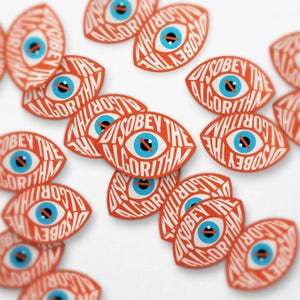 May include: Orange eye-shaped stickers with the text "DISOBEY THE ALGORITHM" in white. The eye has a blue iris and a black line through it. The stickers are scattered on a white surface.