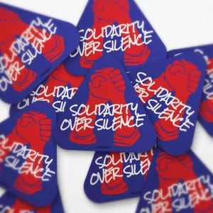 May include: A collection of blue, triangular stickers with the text "SOLIDARITY OVER SILENCE" in white. Each sticker features a red fist graphic. The stickers are scattered, creating a layered effect.