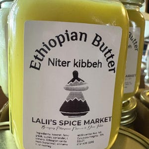 Op de afbeelding: Een glazen pot gevuld met gele Ethiopische boter, met het label "Ethiopian Butter Niter kibbeh" en "Lalii's Spice Market". Het label bevat ook de ingrediënten, het adres en het telefoonnummer van de specerijenmarkt. De pot is 16 ounce of 453,6 gram.