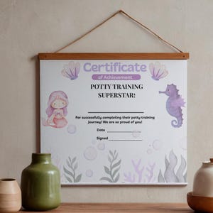 May include: A framed certificate with a wooden hanger, featuring a mermaid and seahorse illustration. The certificate reads "Potty Training Superstar!" in purple and black text, with space for a date and signature. The background has a light purple and gray underwater theme.