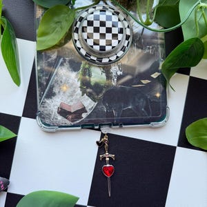 May include: A clear phone case featuring a collage of a dark wolf, an open book, and a moon, topped with a black and white chequered circular phone grip. A silver-toned sword charm with a red heart-shaped gemstone and a small black heart hangs from the bottom.
