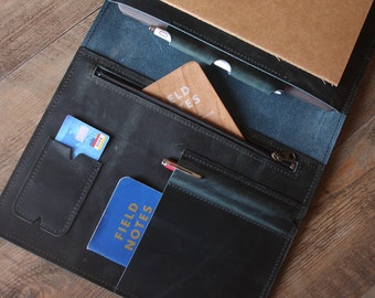 May include: A dark blue leather portfolio with a zippered pocket, a credit card slot, and two pen holders. The portfolio is open and contains a notebook, a pen, and a wooden bookmark with the text "Field Notes".