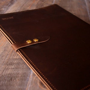 May include: A brown leather portfolio with a flap closure and two brass rivets. The portfolio has a stitched edge and the text "Just Craft" is embossed on the front.
