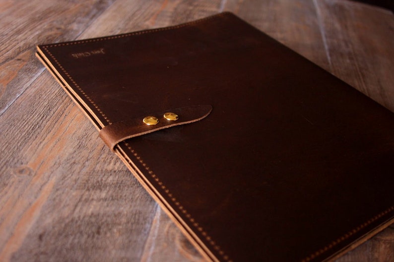 May include: A brown leather portfolio with a flap closure and two brass rivets. The portfolio has a stitched edge and the text "Just Craft" is embossed on the front.