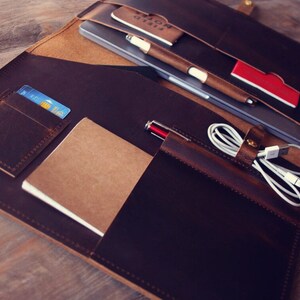 May include: A brown leather portfolio with multiple pockets and compartments. It contains a laptop, a notebook, a pen, a credit card, and a charging cable.