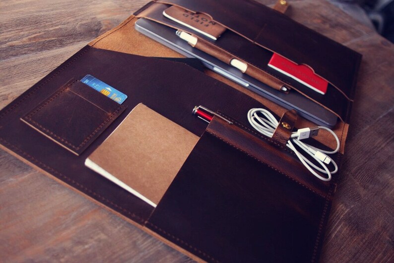 May include: A brown leather portfolio with multiple pockets and compartments. It contains a laptop, a notebook, a pen, a credit card, and a charging cable.