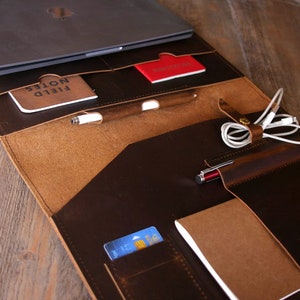 May include: A brown leather laptop portfolio with multiple pockets and compartments. The portfolio is open and contains a laptop, a notebook, a pen, a credit card, and a pair of headphones.