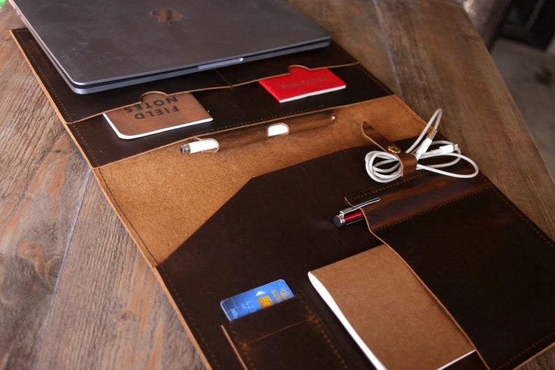 May include: A brown leather laptop portfolio with multiple pockets and compartments. The portfolio is open and contains a laptop, a notebook, a pen, a credit card, and a pair of headphones.