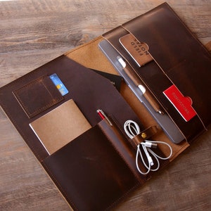 May include: A brown leather portfolio with multiple pockets and compartments. The portfolio is open and contains a tablet, a notebook, a pen, a credit card, and a charging cable. The portfolio is on a wooden surface.