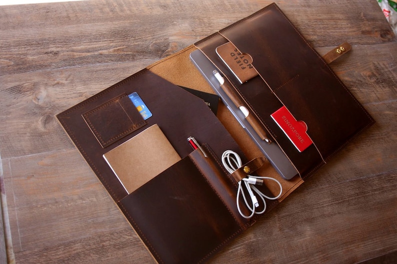 May include: A brown leather portfolio with multiple pockets and compartments. The portfolio is open and contains a tablet, a notebook, a pen, a credit card, and a charging cable. The portfolio is on a wooden surface.