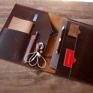 Könnte beinhalten: Eine braune Ledertasche für Laptops mit mehreren Taschen und Fächern. Die Tasche ist geöffnet und zeigt einen Laptop, einen Stift, einen Notizblock, eine Kreditkarte und Kopfhörer.