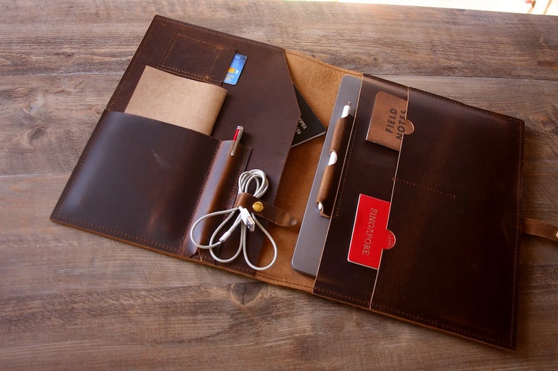 May include: A brown leather laptop sleeve with multiple pockets and compartments. The sleeve is open and shows a laptop, a pen, a notepad, a credit card, and a pair of headphones.