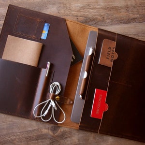 May include: A brown leather portfolio with multiple pockets and compartments. The portfolio is open and contains a laptop, a notebook, a pen, and a few cards. The cards have the text "Field Notes" and "Singapore" on them.