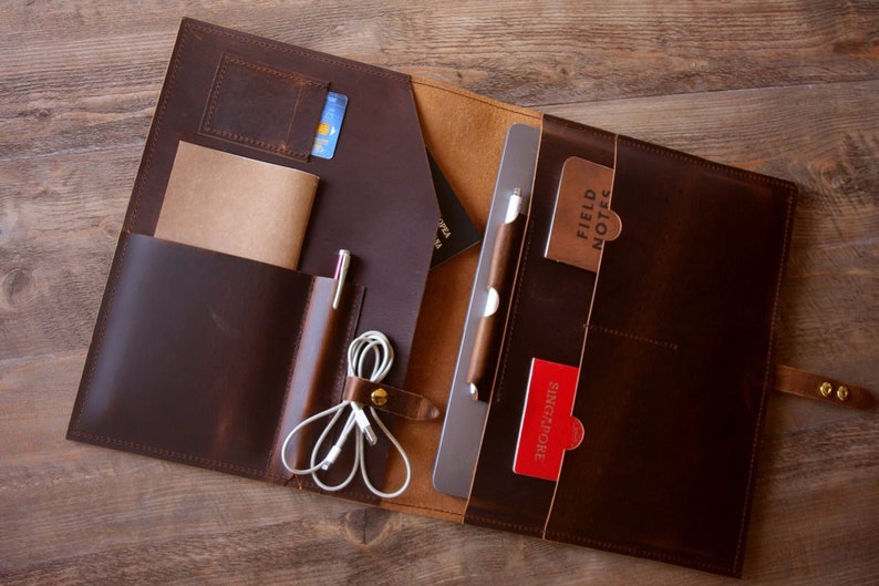 May include: A brown leather portfolio with multiple pockets and compartments. The portfolio is open and contains a laptop, a notebook, a pen, and a few cards. The cards have the text "Field Notes" and "Singapore" on them.