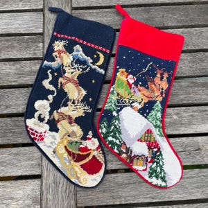 Puede incluir: Dos calcetines navideños con diseños bordados. Uno es azul marino con Papá Noel y renos, el otro es rojo y azul marino con una escena similar. Ambos tienen un lazo para colgar y están sobre una superficie de madera.