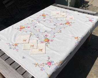 May include: A white rectangular tablecloth and four matching square napkins featuring embroidered floral motifs in pink, yellow, and blue. The central design includes a decorative border pattern with intricate stitching and colorful flower clusters.