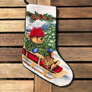 May include: A white Christmas stocking with a green, red, and brown embroidered design of a teddy bear wearing a red hat and blue mittens riding a red sled in a snowy forest.