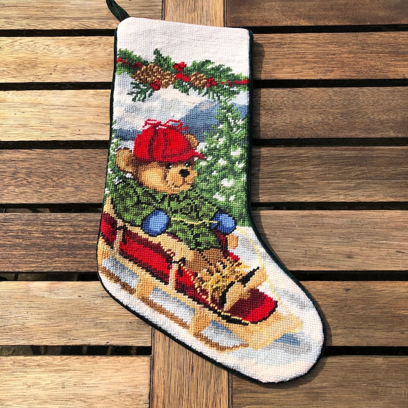 May include: A white Christmas stocking with a green, red, and brown embroidered design of a teddy bear wearing a red hat and blue mittens riding a red sled in a snowy forest.