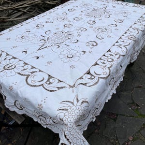 Pode incluir: Toalha de mesa retangular branca com um design floral e abacaxi detalhado em marrom. As bordas apresentam uma borda ondulada. A toalha de mesa é colocada sobre uma mesa de metal escuro, com um fundo natural ao ar livre.