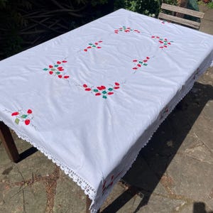 以下が含まれることがあります： 赤と緑の刺繍の花柄が施された白い長方形のテーブルクロス。 テーブルクロスの端には繊細なレースのトリムが付いています。 刺繍された花は、テーブルクロスの中心に長方形のパターンで配置されています。