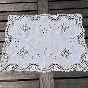 Peut inclure: Nappe rectangulaire blanche avec une bordure en dentelle délicate et des motifs ajourés complexes. Le centre présente des motifs en forme de losange et des accents floraux, créant une esthétique élégante et vintage. La nappe est posée sur une surface en bois.