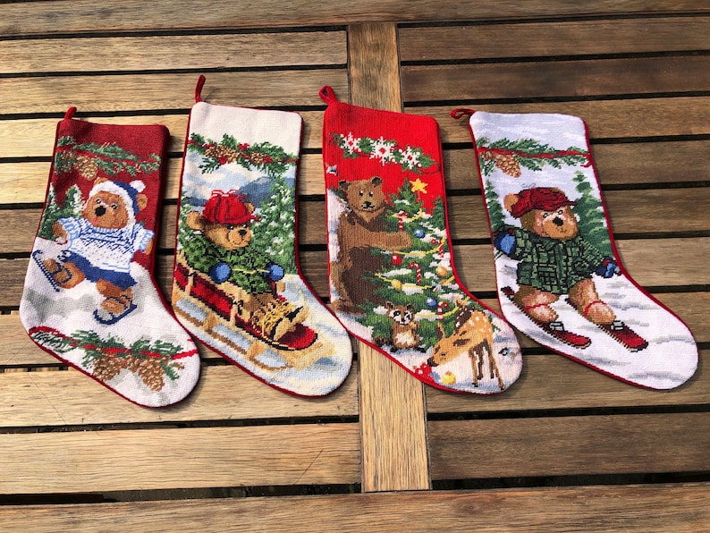 May include: Four red and white Christmas stockings with embroidered teddy bears and winter scenes. The stockings feature a bear ice skating, a bear on a sled, a bear decorating a Christmas tree, and a bear skiing.