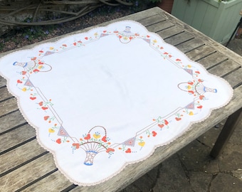 Puede incluir: Mantel blanco con borde festoneado y diseño floral bordado con cestas de flores en tonos naranja, rojo, amarillo y azul.