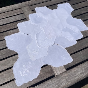 Può includere: Una collezione di centrini bianchi con bordi smerlati e delicati ricami floreali. I centrini sono disposti in un motivo a strati, mostrando i loro intricati disegni. L'immagine è scattata all'aperto su una superficie di legno, evidenziando l'estetica vintage.