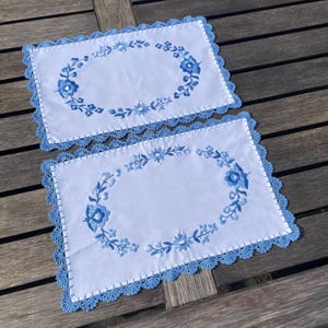 Op de afbeelding: Twee witte gehaakte placemats met blauwe geborduurde bloemenmotieven. De placemats hebben een gegolfde rand.