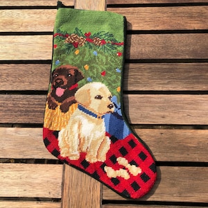May include: A green Christmas stocking with a red and black plaid cuff features two dogs, a brown Labrador Retriever and a golden retriever, sitting in front of a Christmas tree with presents.