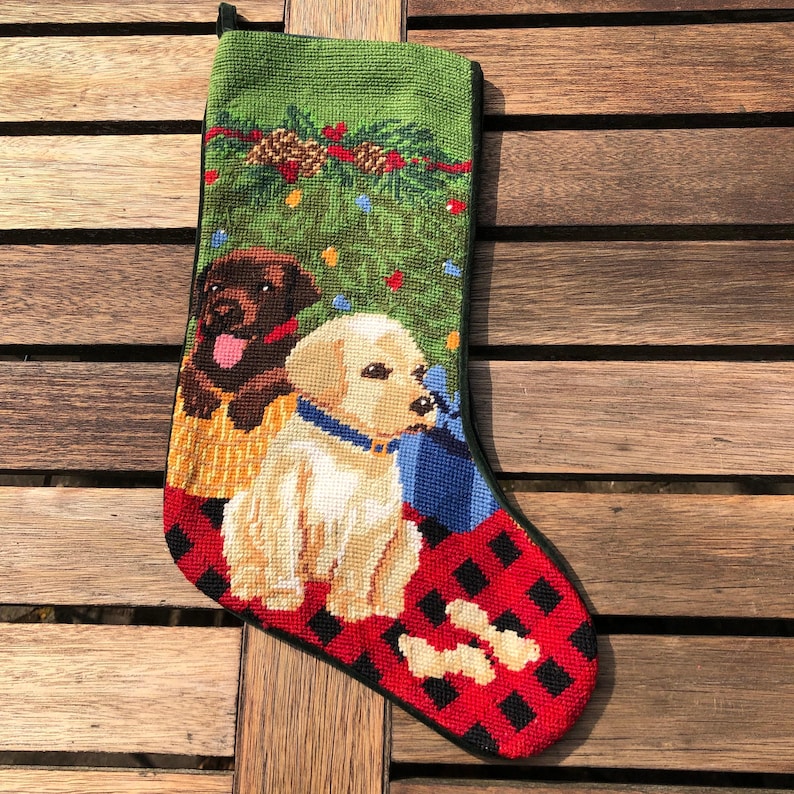 May include: A green Christmas stocking with a red and black plaid cuff features two dogs, a brown Labrador Retriever and a golden retriever, sitting in front of a Christmas tree with presents.
