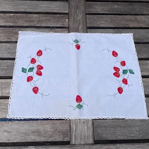May include: A rectangular white placemat featuring embroidered red strawberries with green leaves arranged in two curved side patterns and two single centred motifs, finished with a delicate white crocheted lace border along all four edges.