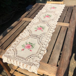 May include: Cream-colored table runner with embroidered pink roses and green leaves. The runner has a crocheted lace border. The runner is placed on a wooden table.