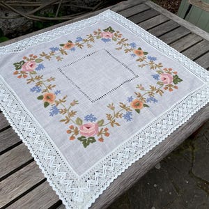 以下が含まれることがあります： ピンク、ブルー、ブラウンの色合いの刺繍された花柄が施された、繊細なレースの縁取りが施された白いリネン製のテーブルクロスです。