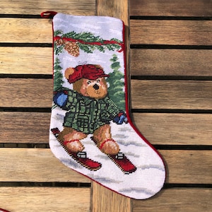May include: A white Christmas stocking with a red border features a needlepoint design of a brown bear wearing a green plaid shirt and red hat, skiing down a snowy hill.