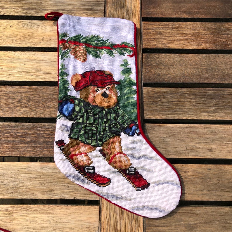 May include: A white Christmas stocking with a red border features a needlepoint design of a brown bear wearing a green plaid shirt and red hat, skiing down a snowy hill.