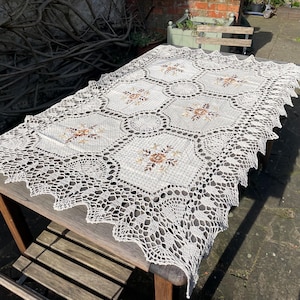 Puede incluir: Un mantel de crochet blanco con un delicado patrón floral. El mantel tiene un borde festoneado y está colocado sobre una mesa de madera.