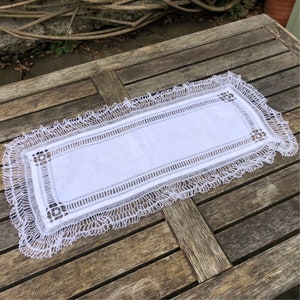 May include: White crocheted table runner with a delicate lace border. The runner is laid out on a wooden table.