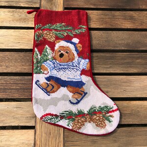 May include: Red and white Christmas stocking with a brown bear wearing a blue hat and sweater ice skating on a white background. The stocking is decorated with green pine branches and brown pine cones.