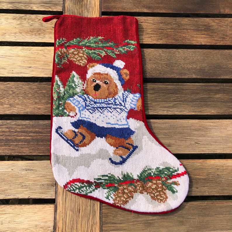 May include: Red and white Christmas stocking with a brown bear wearing a blue hat and sweater ice skating on a white background. The stocking is decorated with green pine branches and brown pine cones.