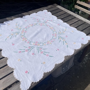 以下が含まれることがあります： 中央に刺繍された花のリースと、ピンク、黄色、緑の色合いで散りばめられた花の小枝が特徴の正方形の白いテーブルクロス。生地の縁はスカラップ状になっており、周囲全体に繊細な刺繍が施されています。