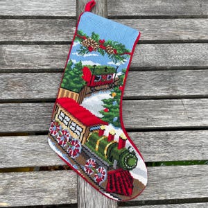 May include: Needlepoint Christmas stocking with a red trim and loop. The stocking features a detailed scene of a green train, a red train, and a snowy landscape with pine trees and a blue sky. The stocking is approximately 46 cm long.