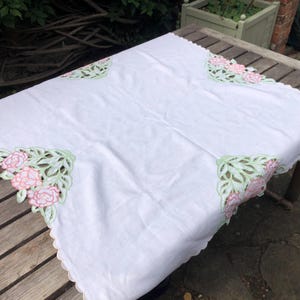 Puede incluir: Un mantel rectangular blanco con bordes festoneados y un intrincado bordado calado en las esquinas. El bordado muestra motivos florales estilizados en tonos rosa suave y verde claro, creando un diseño de borde decorativo.
