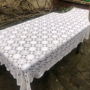 May include: White crocheted tablecloth with a delicate floral pattern. The tablecloth is laid out on a wooden table.
